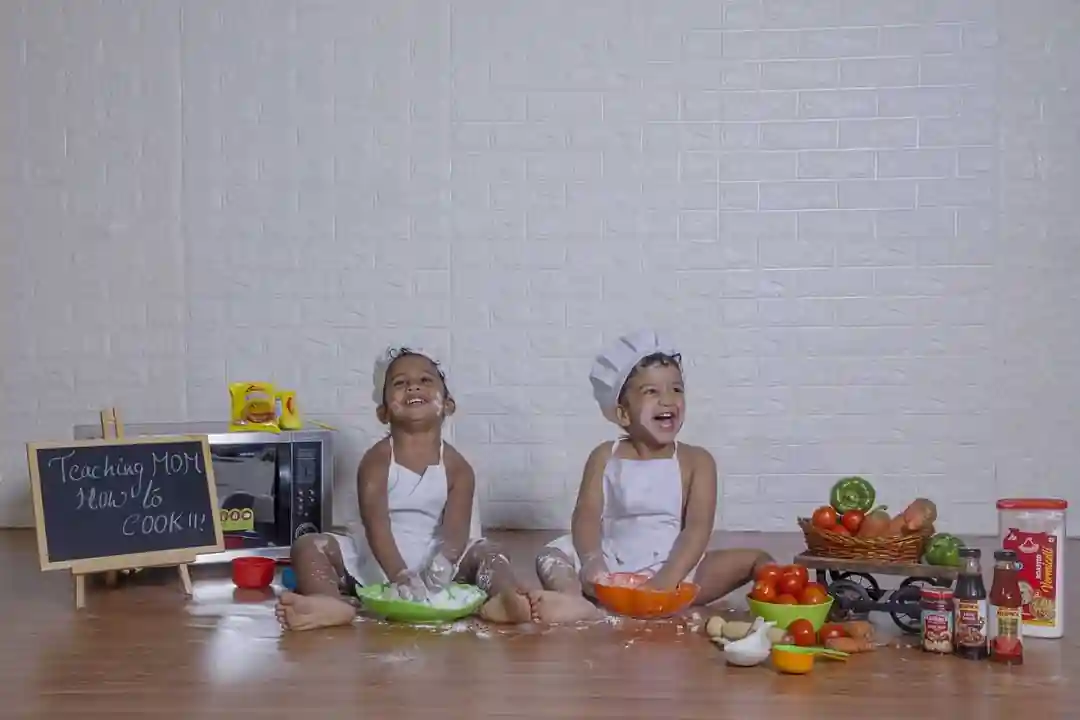 Two kids in chef outfits having fun in the kitchen, with flour on their faces, surrounded by cooking ingredients and a chalkboard sign that says 'Teaching MOM how to cook'.