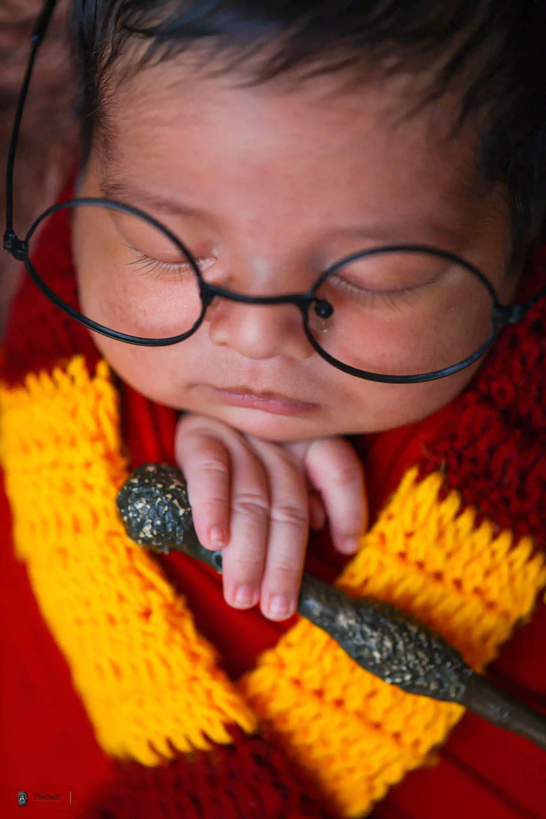 Newborn baby dressed in Harry Potter-themed outfit with glasses and wand, sleeping peacefully.