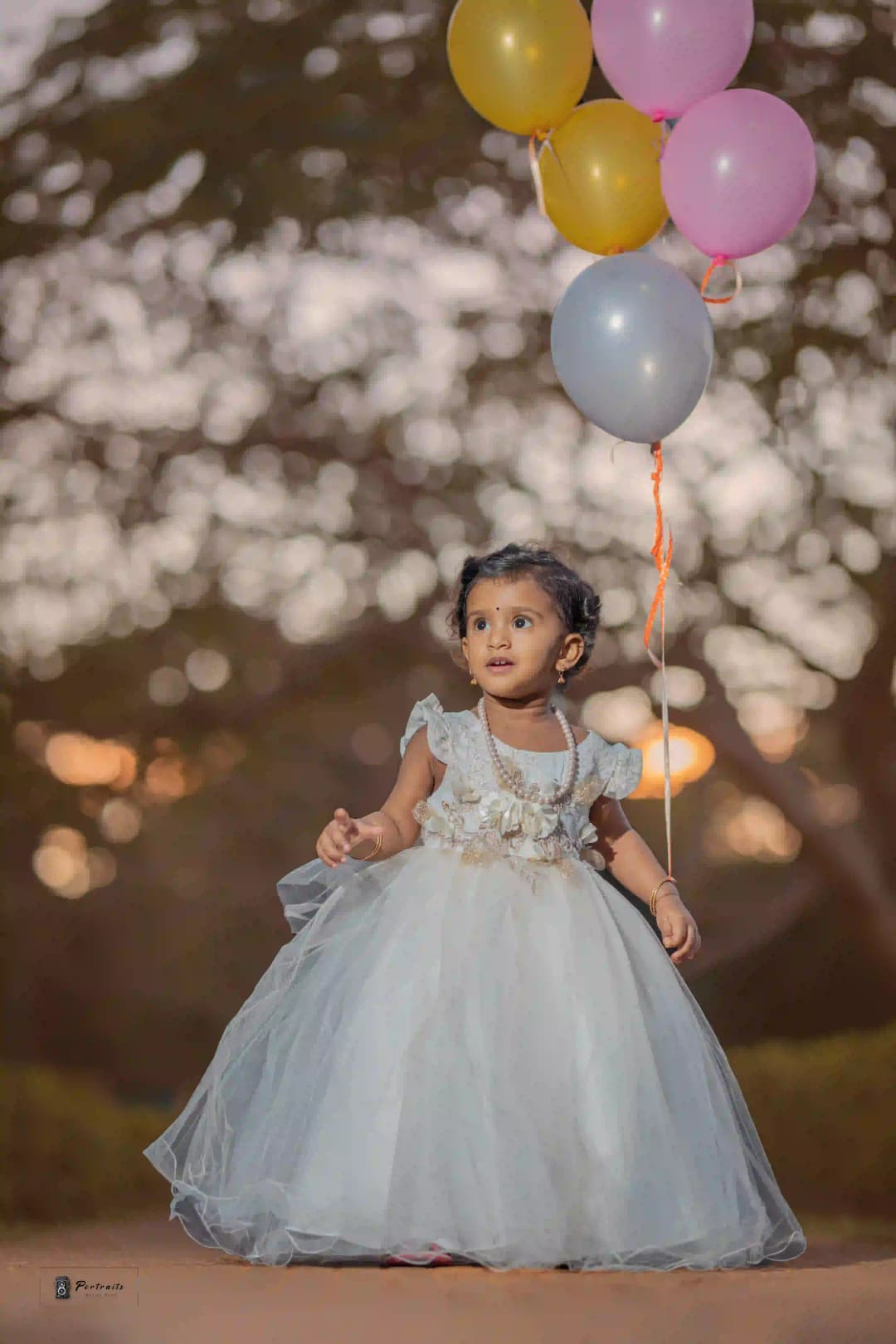 Happy toddler in a white dress holding colorful balloons outdoors during sunset.