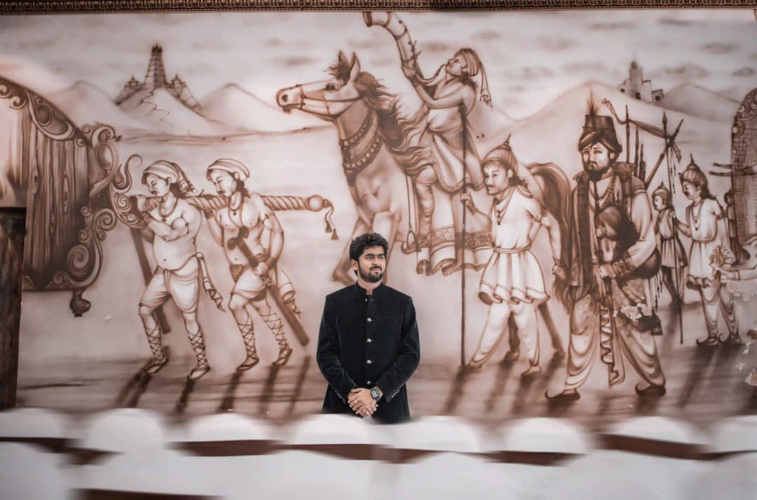 Man standing in front of a large historical mural depicting ancient warriors and soldiers, with a traditional attire.