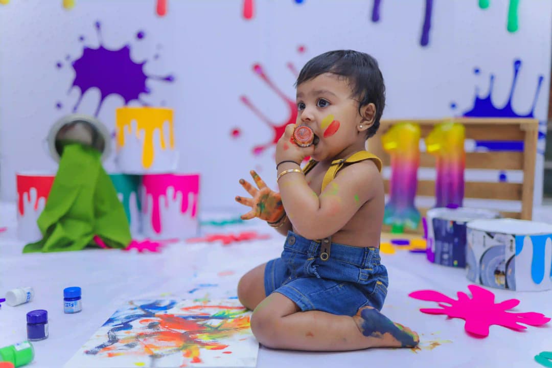 Baby playing with colorful paints, sitting on the floor with splashes of paint around, holding a paintbrush, and creating a colorful mess.