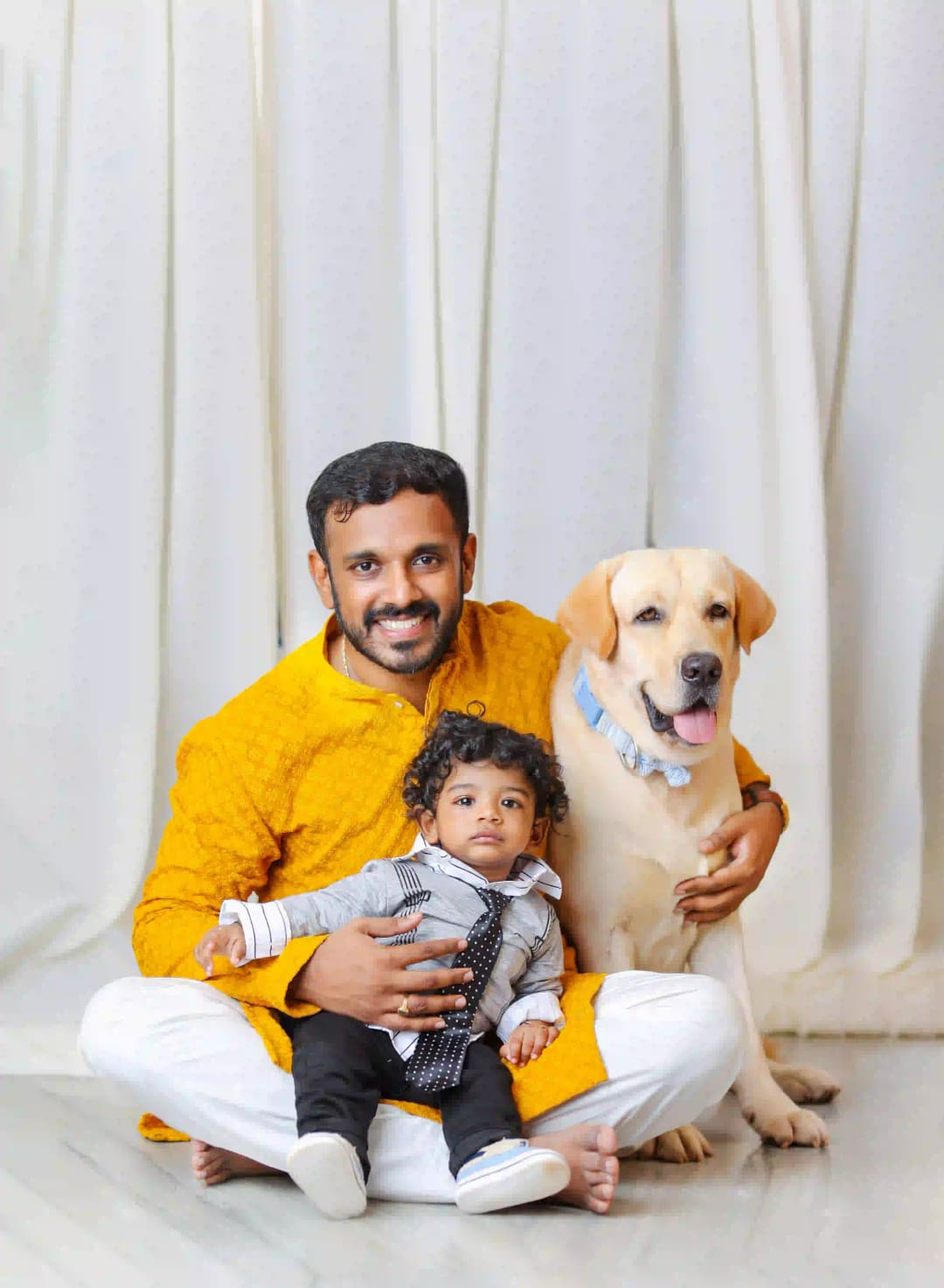 A happy father with his toddler and Labrador dog, dressed in traditional attire, smiling for a family portrait.