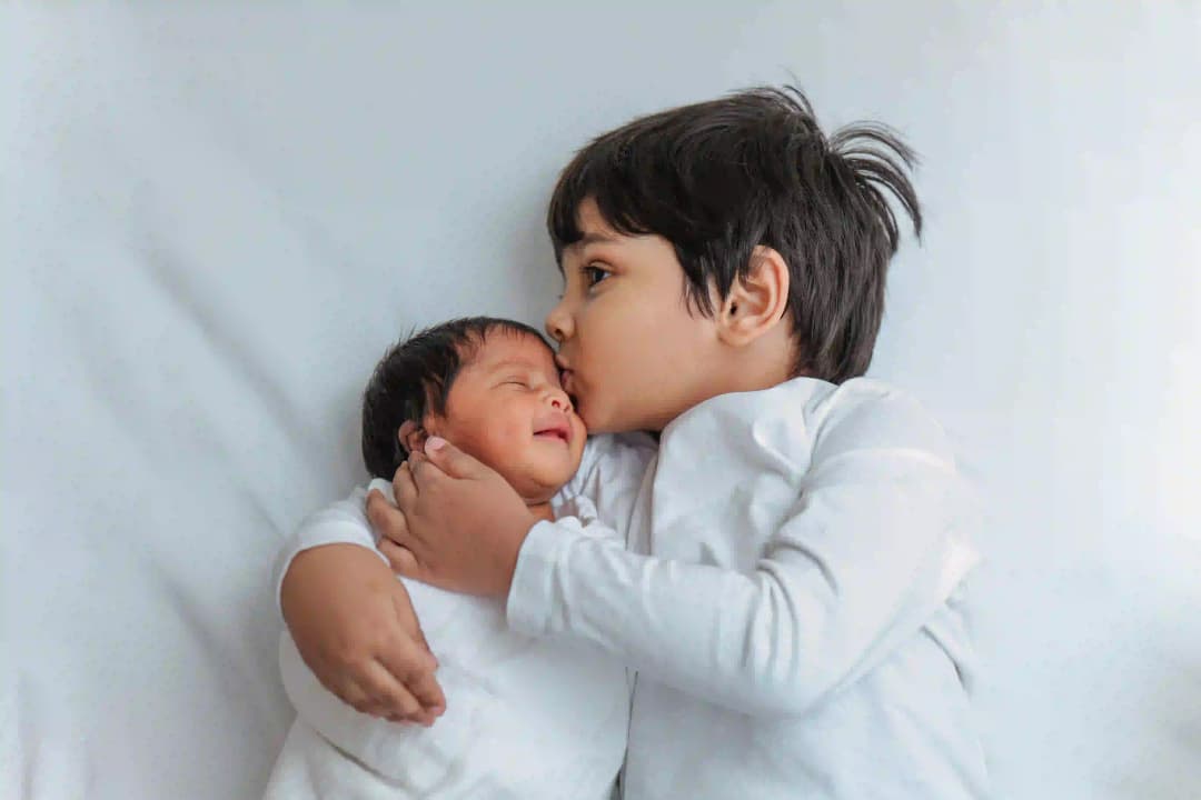 Older sibling lovingly kisses their newborn baby brother or sister, capturing a tender sibling moment