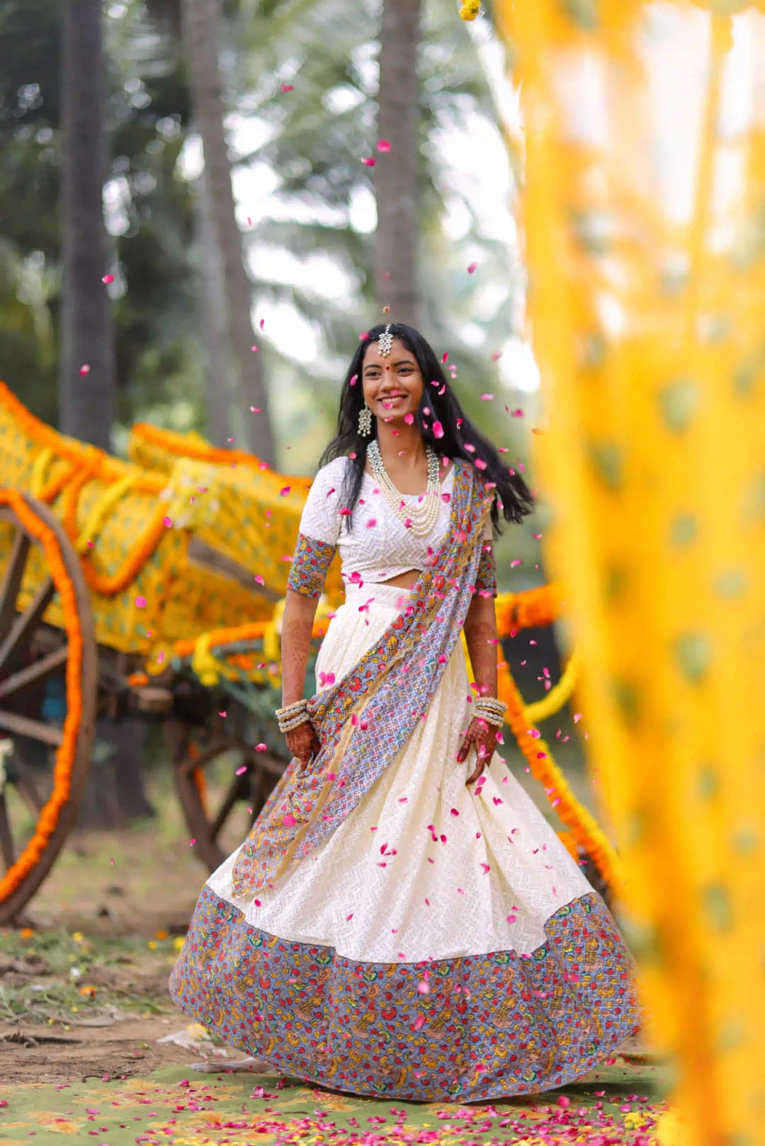 Woman in traditional white attire with floral embroidery, walking amidst flower petals during a cultural celebration.
