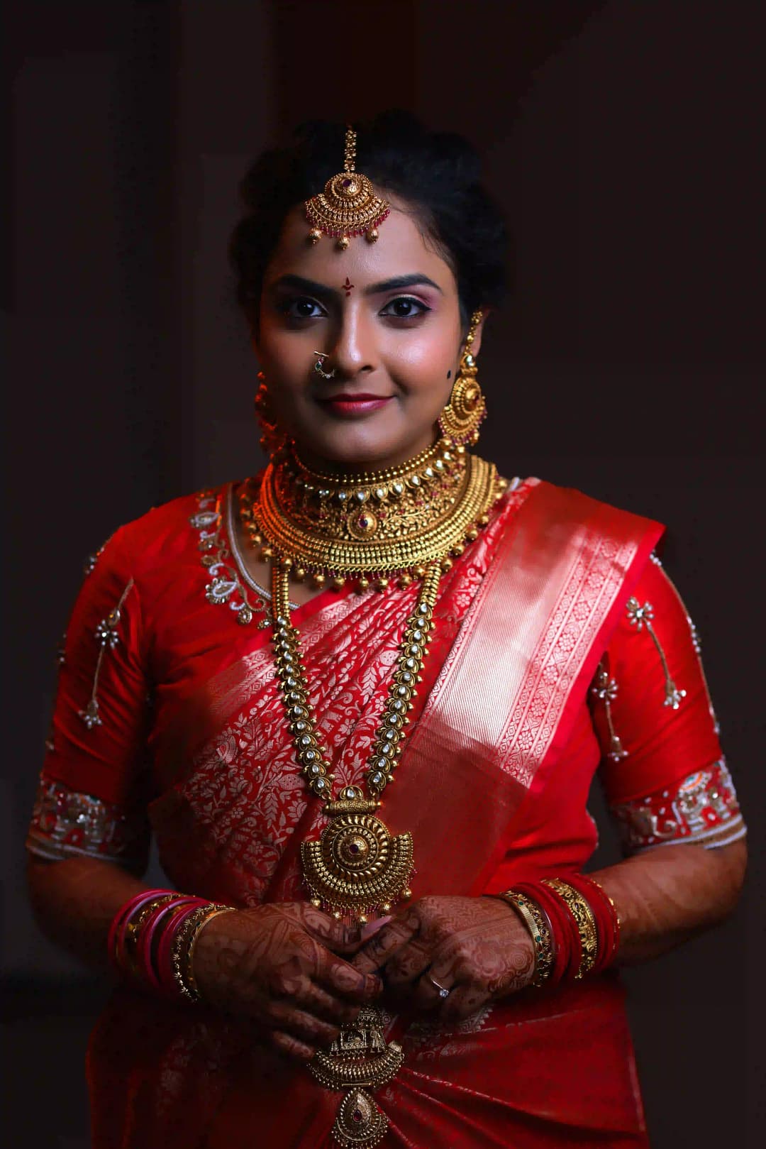 Portrait of a woman in a red bridal saree adorned with gold jewelry, including necklaces, bangles, and a nose ring.